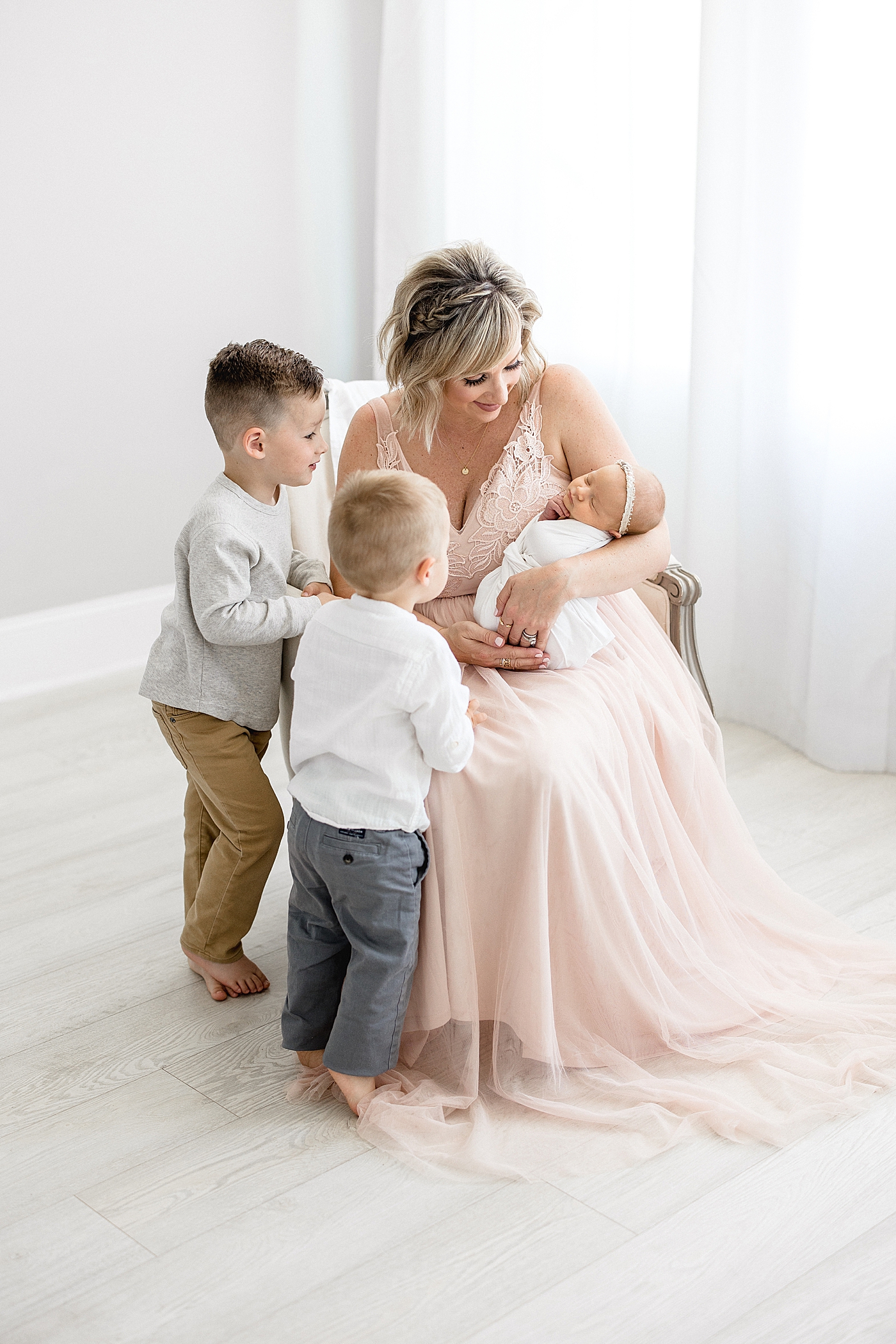 Mom sitting in chair with baby and big brothers are looking at her. Photo by Brittany Elise Photography.