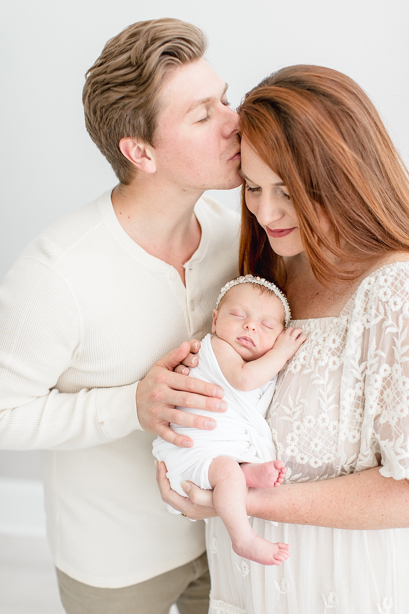 Dad kissing Mom on the. forehead while she holds their newborn daughter. Photo by Brittany Elise Photography.
