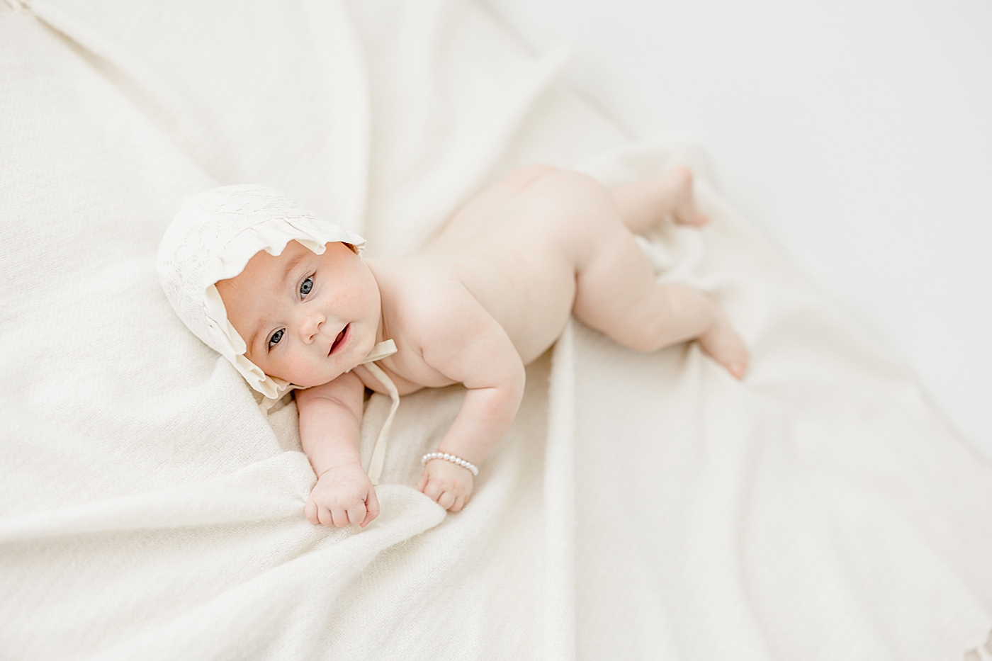 Six month old baby girl laying on her side with a sweet bonnet and pearl bracelet on. Photo by Brittany Elise Photography.