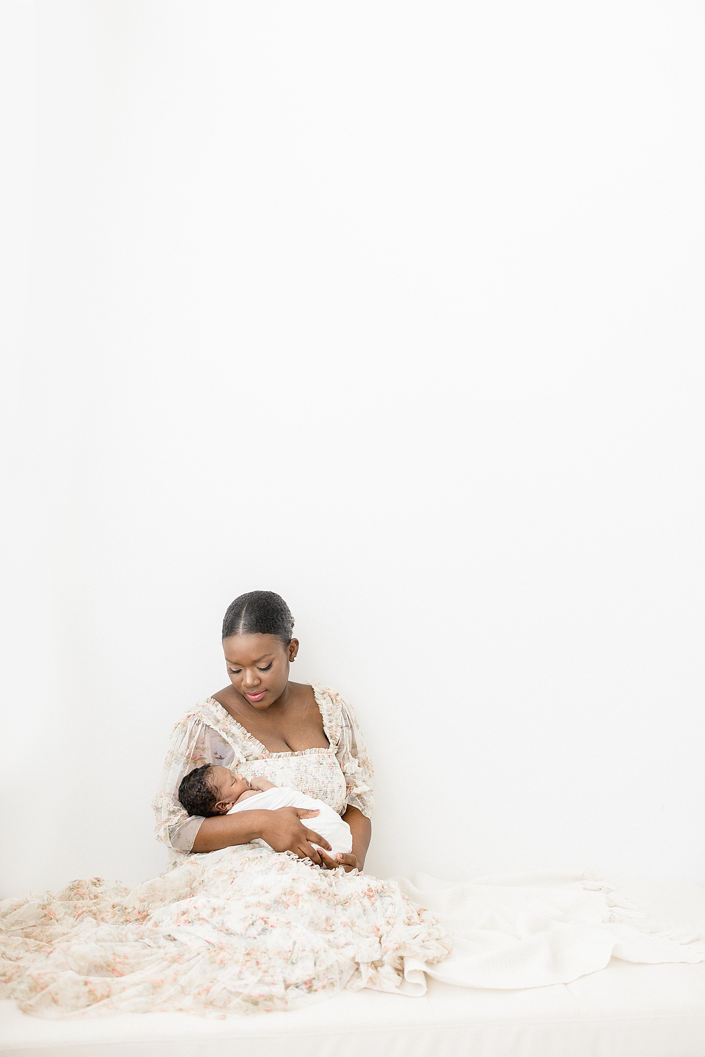 Mom wearing floral dress sitting on bed holding her newborn son. Photo by Brittany Elise Photography.